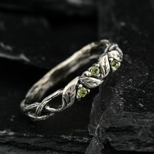 May include: A silver ring featuring a textured band designed to resemble intertwined branches and leaves. The band is accented with three small, round, light green gemstones set within the metal foliage, creating a detailed botanical appearance.