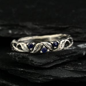 May include: A silver ring featuring a band sculpted with a detailed leaf and vine pattern. Three small, round, deep blue gemstones are set at intervals along the top portion of the band, integrated into the organic, textured metal design.