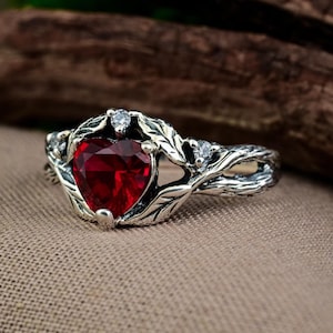 Könnte beinhalten: Silberner Ring mit einem herzförmigen, facettierten roten Edelstein. Der Ring hat ein Blatt- und Weinreben-Design mit kleinen, klaren Akzentsteinen. Der Ring ist vor einem neutralen Hintergrund platziert.