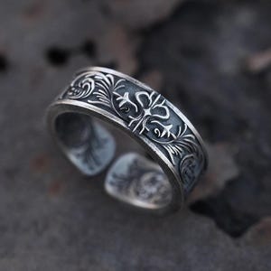 Op de afbeelding: Een zilveren verstelbare ring met een sierlijk bloemmotief. De ring heeft gedetailleerde gravures van wervelende bladeren en symmetrische patronen. Het open ontwerp maakt maatverstelling mogelijk. De ring wordt gepresenteerd op een donkere, getextureerde ondergrond.