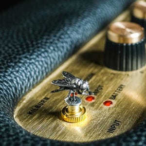 May include: Close-up of a black and gold Marshall Kilburn speaker. A detailed silver fly-shaped knob is featured on the control panel. The panel has the word "INPUT" and "BATTERY" printed on it. The speaker is surrounded by black leather.