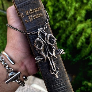 May include: A silver necklace with a detailed pendant featuring two ram skulls with horns. The pendant is hanging on a worn leather-bound book with the text "Al. Testament u. Psalmen".