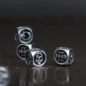 Pode incluir: Cinco dados de metal prateado com desenhos de caveiras em cada face. Os dados estão espalhados sobre uma superfície cinza escura.