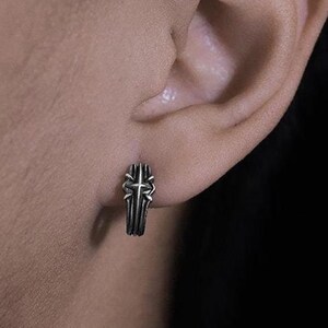 May include: A small silver-toned hoop earring featuring a raised cross design with oxidized detailing. The metal has a textured, weathered finish, and the earring is secured through the earlobe, showcasing a compact and detailed metallic structure.