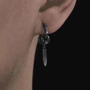 May include: A single silver earring with a dagger pendant. The earring is attached to the ear with a small hoop. The dagger is a long, thin blade with a pointed tip. The earring is worn on the ear.