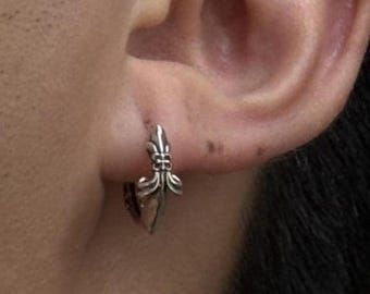 May include: A small silver hoop earring featuring a detailed fleur-de-lis emblem on the front. The earring is secured through the earlobe, showcasing a polished metallic finish with intricate carved lines and a pointed decorative design element.