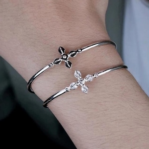Może przedstawiać: Dwie cienkie bransoletki typu bangle w kolorze srebrnym, każda z centralnym ornamentem w kształcie kwiatu lub krzyża o czterech płatkach. Jeden ornament jest z gładkiego polerowanego metalu, a drugi wysadzany małymi, jasnymi kamieniami.