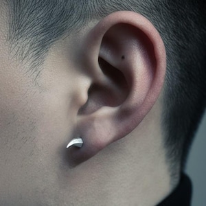 May include: A silver claw-shaped earring is worn in the ear. The earring is a small, curved design. The ear is set against a blurred background. The person is wearing a black turtleneck.
