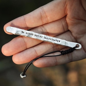 May include: A silver-colored metal bar with the stamped text "TO SHINE NOT BE ILLUMINATED" is held in a hand. A black cord with a small silver bead is attached to the bar.