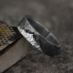 Puede incluir: Un anillo de metal gris oscuro con un acabado texturizado y martillado. La superficie exterior presenta un relieve de cadena montañosa de color plateado pulido y elevado que envuelve parte de la banda, contrastando con el metal oscuro, mate y oxidado.
