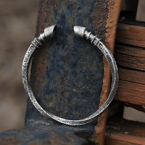 May include: A silver cuff bracelet with an open design. The bracelet has detailed patterns and textured ends. The metal has a slightly aged appearance, suggesting a vintage or handcrafted style. The bracelet is displayed on a weathered, dark surface.