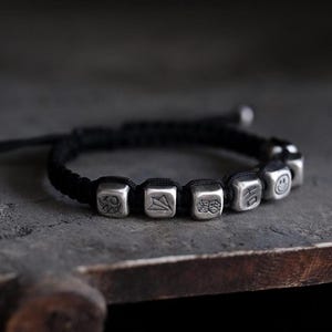 May include: A black cord bracelet with silver cube charms. Each cube has a different symbol engraved on it.