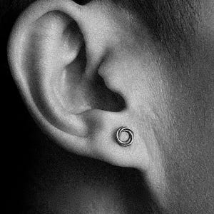 May include: A close-up, black and white image of an ear with a small, silver-colored stud earring. The earring features a circular, intertwined design. The ear is in focus, with the earring centered in the earlobe.