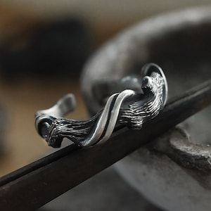 Puede incluir: Un brazalete plateado con un diseño orgánico y texturizado. El brazalete presenta una forma esculpida y fluida con una textura detallada similar a la corteza. El diseño abierto del brazalete es visible.