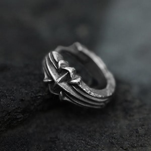 May include: A silver ring with a dark, oxidized finish featuring a prominent raised cross design on the band. The metal surface displays intricate, sculpted detailing with deep grooves and a weathered, textured appearance throughout the entire piece.