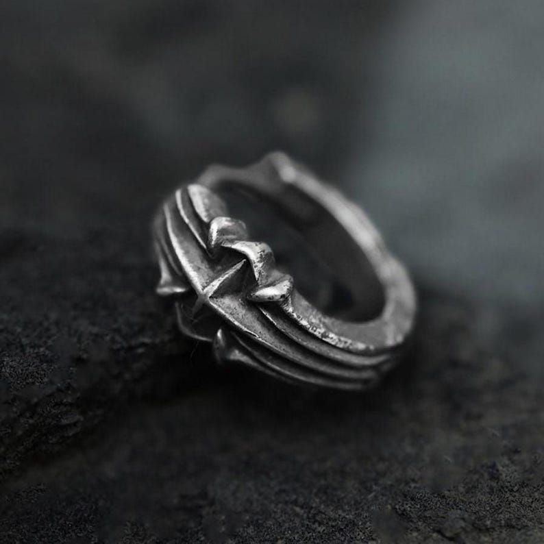 May include: A silver ring with a dark, oxidized finish featuring a prominent raised cross design on the band. The metal surface displays intricate, sculpted detailing with deep grooves and a weathered, textured appearance throughout the entire piece.