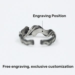 Pu&ograve; includere: Un anello regolabile color argento con un design strutturato e organico. L'anello presenta una forma ondulata e fluida con un'apertura nella parte inferiore. L'immagine include il testo "Engraving Position" e "Free engraving, exclusive customization".