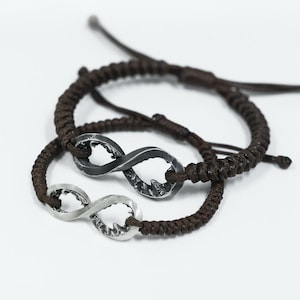 May include: Two matching infinity symbol bracelets. One bracelet features a silver infinity charm, while the other has a dark gray infinity charm. Both charms are attached to a brown braided cord. The bracelets are displayed on a white background.