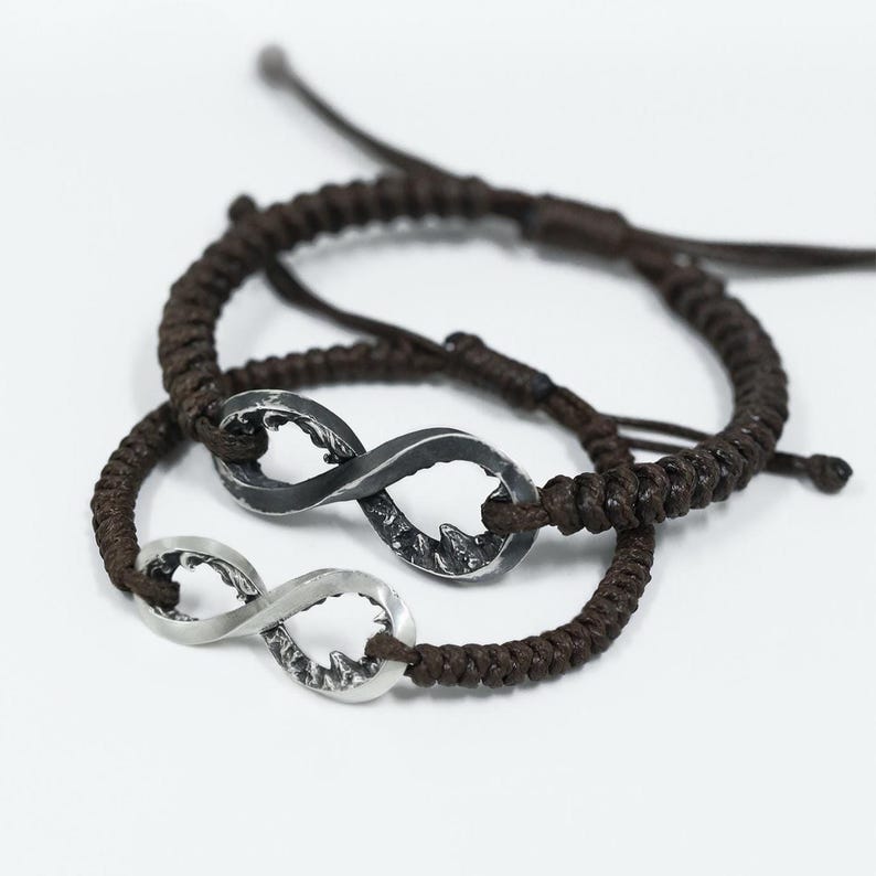 May include: Two matching infinity symbol bracelets. One bracelet features a silver infinity charm, while the other has a dark gray infinity charm. Both charms are attached to a brown braided cord. The bracelets are displayed on a white background.