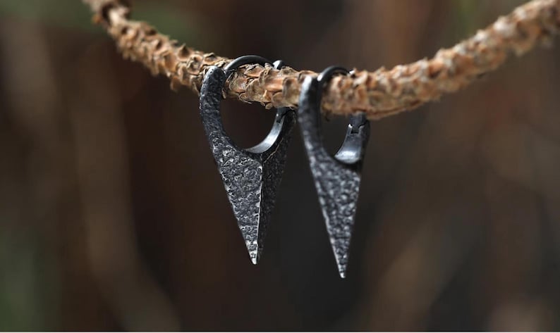 May include: A pair of black metal earrings with a triangular shape and a textured finish. The earrings are hanging from a branch.