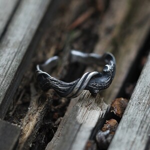 Pu&ograve; includere: Un anello argentato con un design strutturato e organico. L'anello presenta un motivo a forma di ramo attorcigliato, creando un'estetica unica e naturale. L'anello &egrave; appoggiato su una superficie di legno rustico.