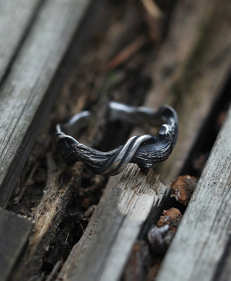 Pu&ograve; includere: Un anello argentato con un design strutturato e organico. L'anello presenta un motivo a forma di ramo attorcigliato, creando un'estetica unica e naturale. L'anello &egrave; appoggiato su una superficie di legno rustico.