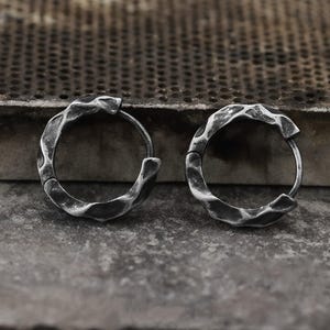 May include: A pair of oxidised silver hoop earrings. Each earring has a textured, hammered design around the hoop. The earrings are displayed on a grey, textured surface.