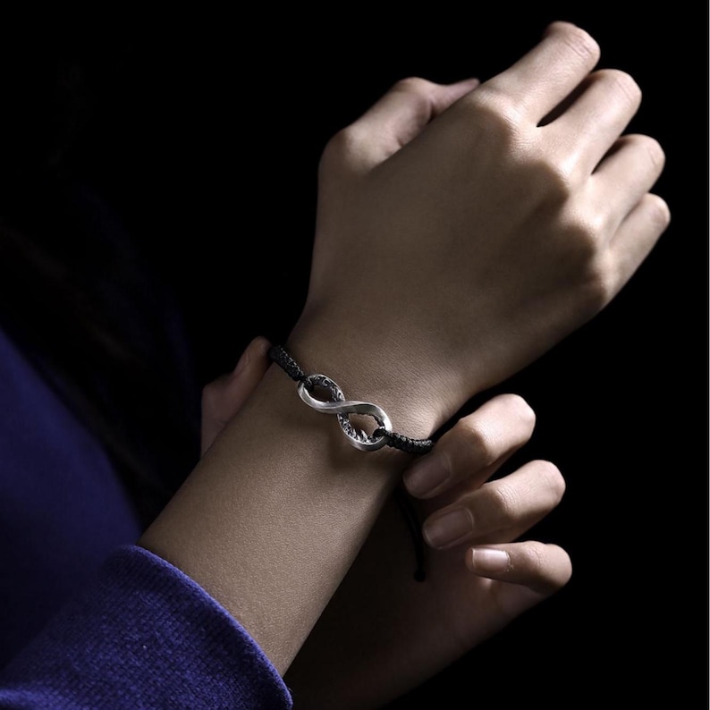 May include: A silver-colored infinity symbol bracelet on a black cord. The bracelet is worn on a wrist, with the hands in the image. The background is dark.