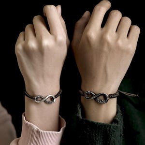 May include: Two matching infinity symbol bracelets. Each bracelet features a silver-colored infinity symbol on a dark brown braided cord. The bracelets are worn on wrists against a black background.