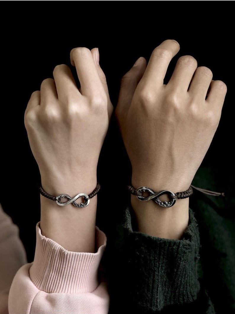 May include: Two matching infinity symbol bracelets. Each bracelet features a silver-colored infinity symbol on a dark brown braided cord. The bracelets are worn on wrists against a black background.