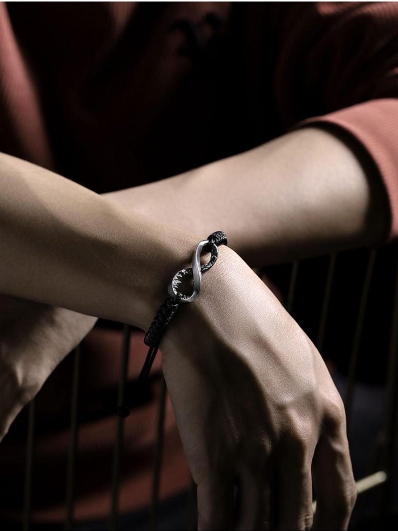 May include: Black braided bracelet featuring a silver infinity symbol. The bracelet has an adjustable cord closure. The infinity symbol is a prominent focal point, adding a touch of elegance to the accessory. The bracelet is designed for everyday wear.