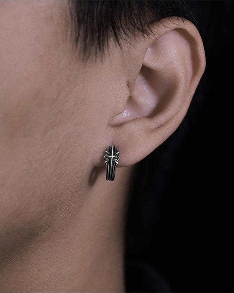 May include: A small silver-toned hoop earring featuring a raised cross design with oxidized detailing. The metal has a textured, weathered finish, and the earring is secured through the earlobe, showcasing a compact and detailed metallic structure.