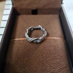 Pu&ograve; includere: Un anello argentato con un design intrecciato e organico, che ricorda rami o viti intrecciate. L'anello &egrave; posto su una superficie marrone strutturata all'interno di una scatola scura, suggerendo un astuccio di presentazione o di conservazione.
