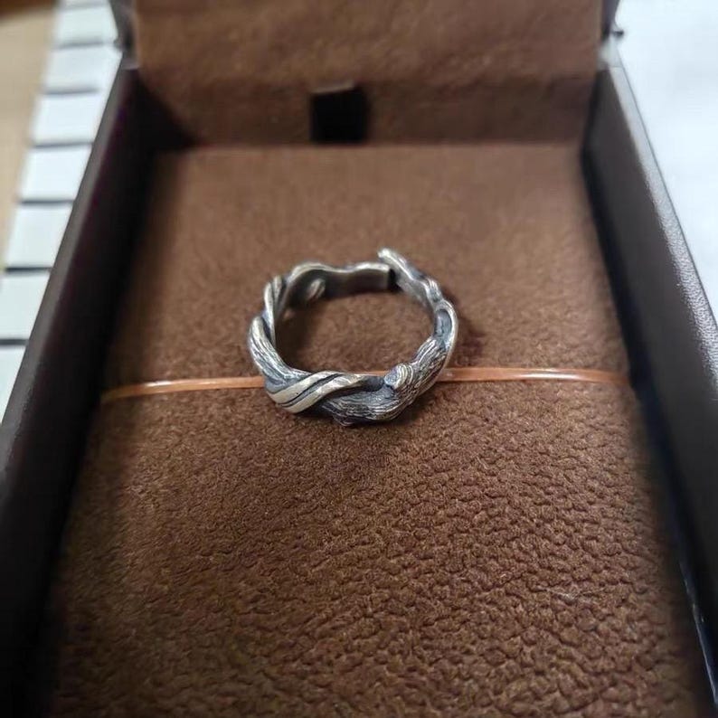 Pu&ograve; includere: Un anello argentato con un design intrecciato e organico, che ricorda rami o viti intrecciate. L'anello &egrave; posto su una superficie marrone strutturata all'interno di una scatola scura, suggerendo un astuccio di presentazione o di conservazione.