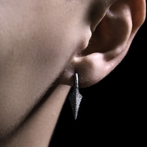 May include: A silver earring with a pointed, textured design. The earring is worn in the ear of a person.