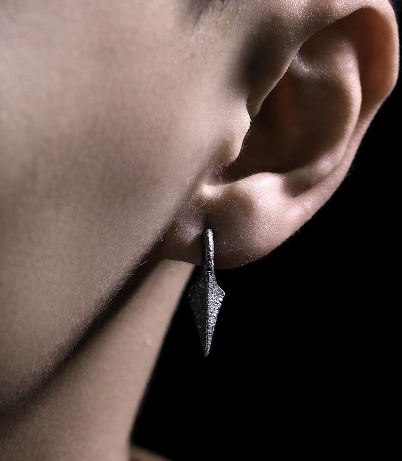 May include: A silver earring with a pointed, textured design. The earring is worn in the ear of a person.