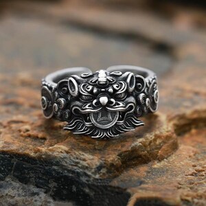 Può includere: Un anello d'argento dettagliato raffigurante il volto di una creatura mitica. L'anello presenta intagli intricati e un design dettagliato del volto della creatura, con un elemento a forma di moneta in bocca. L'anello è posto su una superficie marrone strutturata.