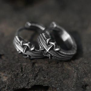 May include: A pair of silver-toned hoop earrings featuring a textured, oxidized finish and a prominent cross-shaped design element on the outer surface. The metal has a weathered appearance with deep grooves and raised details along the circular band.