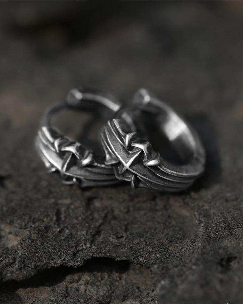 May include: A pair of silver-toned hoop earrings featuring a textured, oxidized finish and a prominent cross-shaped design element on the outer surface. The metal has a weathered appearance with deep grooves and raised details along the circular band.