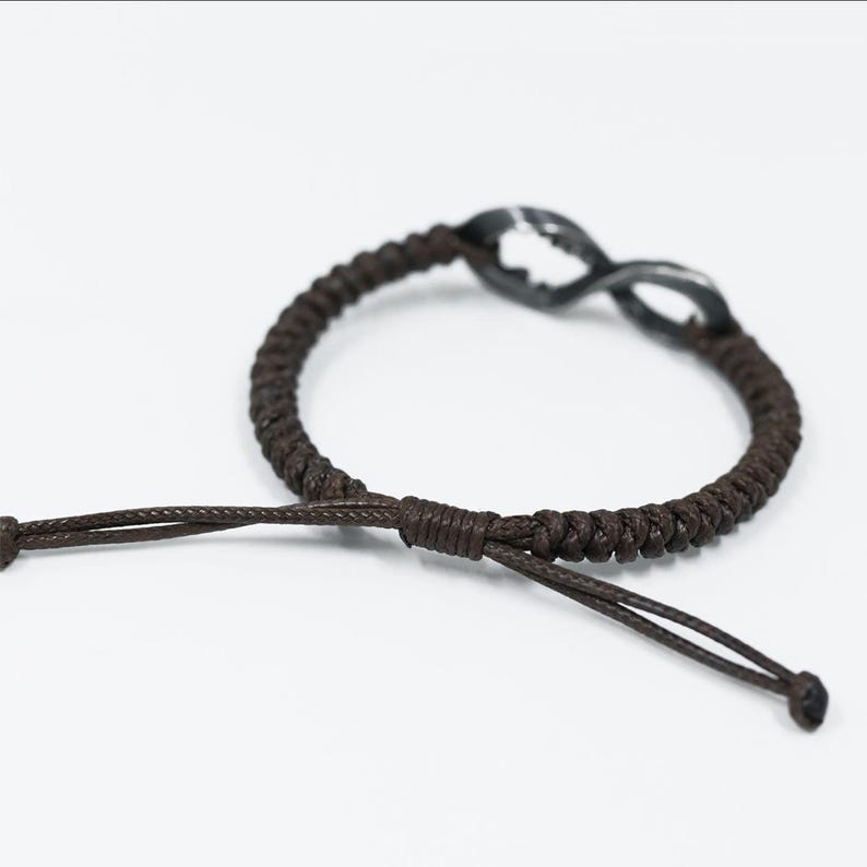 May include: Brown braided bracelet with an infinity symbol charm. The bracelet features a dark brown cord and an adjustable sliding knot closure. The infinity charm is a dark metallic color. The bracelet is displayed on a white background.