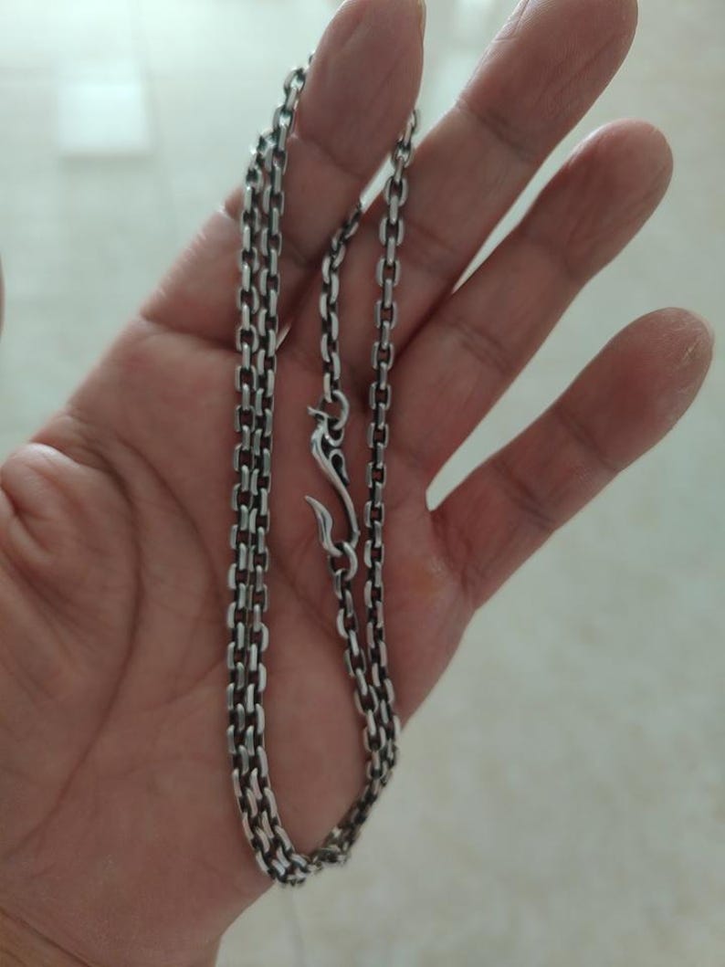 Op de afbeelding: Een zilveren ketting met een karabijn sluiting. De ketting is gemaakt van kleine, met elkaar verbonden vierkanten.