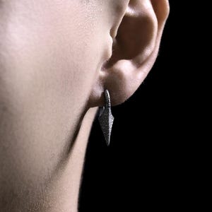 May include: A silver earring with a pointed, triangular design. The earring is worn in the left earlobe.