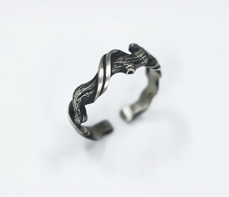 Pu&ograve; includere: Un anello regolabile color argento con un design strutturato e organico. L'anello presenta un motivo attorcigliato e fluido con una texture ruvida simile alla corteccia. Il design aperto consente la regolazione delle dimensioni. L'anello &egrave; su sfondo bianco.