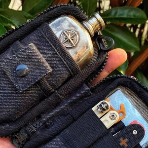 Pu&ograve; includere: Una pochette nera con cerniera con una fiaschetta a forma di bussola e un multi-tool con un design a forma di teschio. La pochette ha una texture simile al denim.