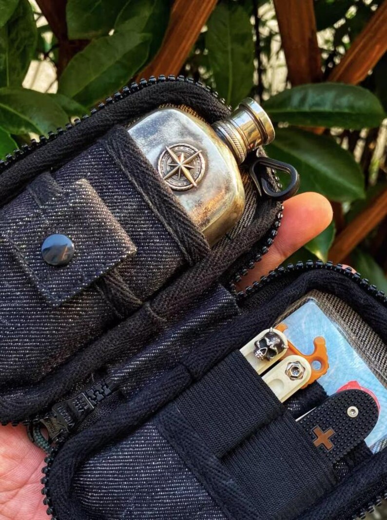 Pu&ograve; includere: Una pochette nera con cerniera con una fiaschetta a forma di bussola e un multi-tool con un design a forma di teschio. La pochette ha una texture simile al denim.