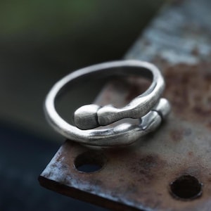 May include: A silver ring with an abstract, flowing design. The ring is made of metal and has a textured surface. The ring is positioned on a rusty metal surface.