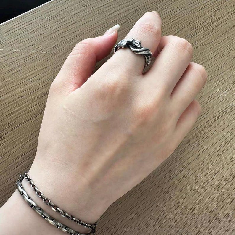 Pu&ograve; includere: Un anello argentato con un design strutturato e intrecciato &egrave; indossato su un dito. Un braccialetto d'argento abbinato con due fili &egrave; visibile sul polso. Lo sfondo &egrave; una superficie di legno di colore chiaro.