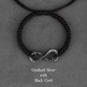 May include: A black cord bracelet featuring an oxidized silver infinity symbol. The bracelet is made of a woven black cord and has the text "Oxidized Silver with Black Cord".