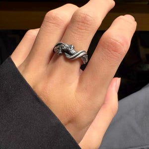 Pu&ograve; includere: Un anello color argento con un design dettagliato ispirato alla natura. L'anello presenta una fascia attorcigliata con un elemento intricato e strutturato, che potrebbe raffigurare un ramo o una forma organica. L'anello &egrave; indossato su un dito, mostrando il suo design e la sua maestria.