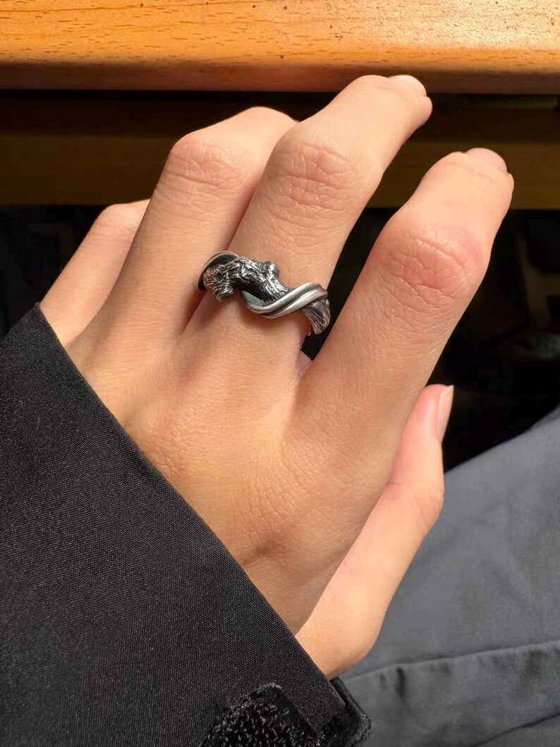 Pu&ograve; includere: Un anello color argento con un design dettagliato ispirato alla natura. L'anello presenta una fascia attorcigliata con un elemento intricato e strutturato, che potrebbe raffigurare un ramo o una forma organica. L'anello &egrave; indossato su un dito, mostrando il suo design e la sua maestria.